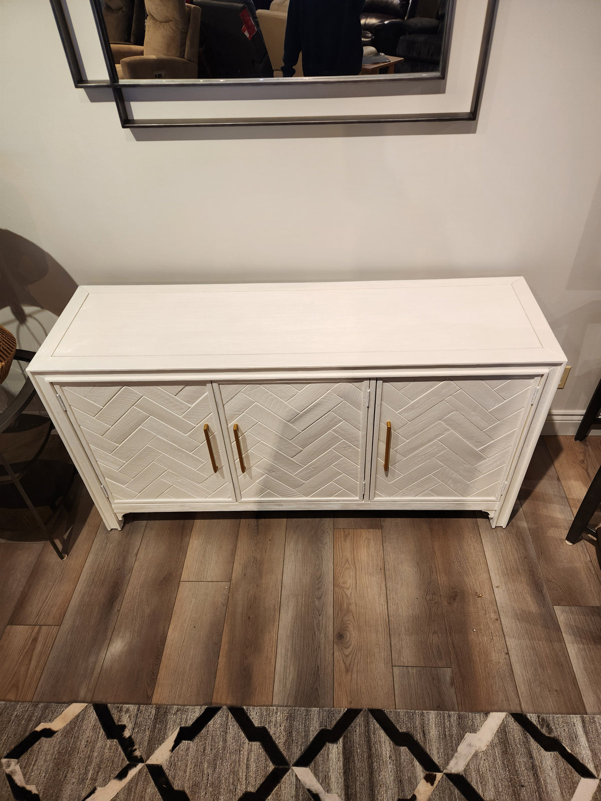 Jofran's Gramercy 3 Door Accent Cabinet with gold handles and chevron inlay stands by a rug and mirror above.