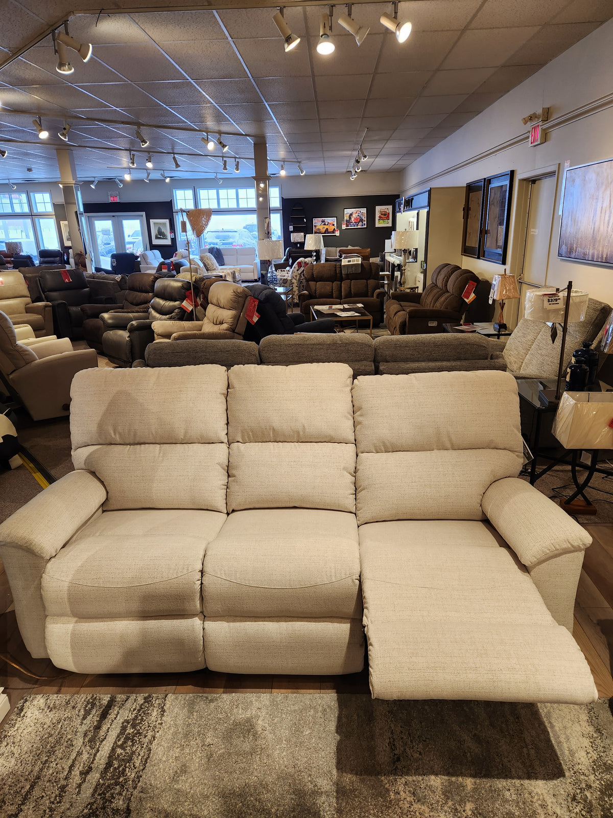 A beige La-z-boy 727 Brooks Reclining Sofa with one footrest extended, shown in a furniture showroom.