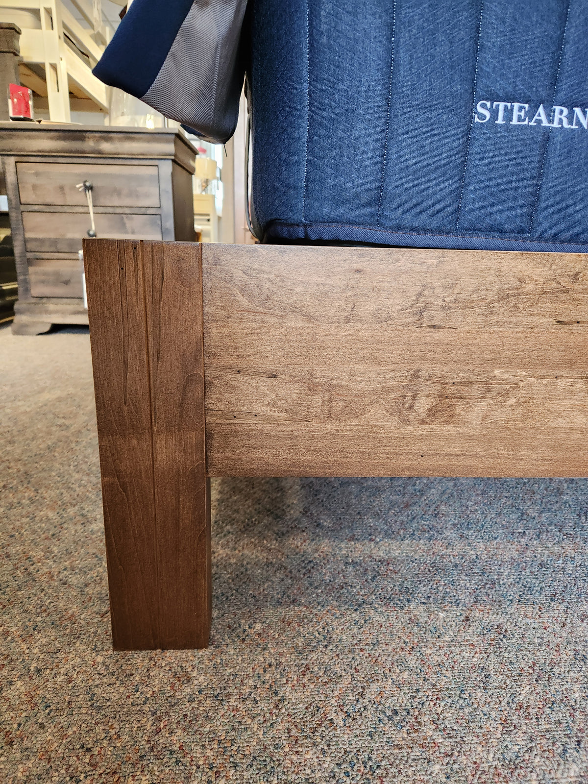 Close-up of the Vokes 247 Vokes Saugeen King Bed Frame leg and rail on carpet, with furniture in the background.
