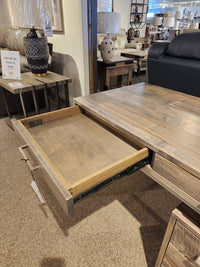 A Handstone EL2448 Electra Writing Desk in driftwood heritage maple with one drawer open sits in a showroom.