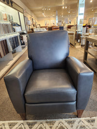 The La-z-boy 431 Scarlett High-Leg Recliner in dark gray leather is showcased in a showroom with fabric samples behind.