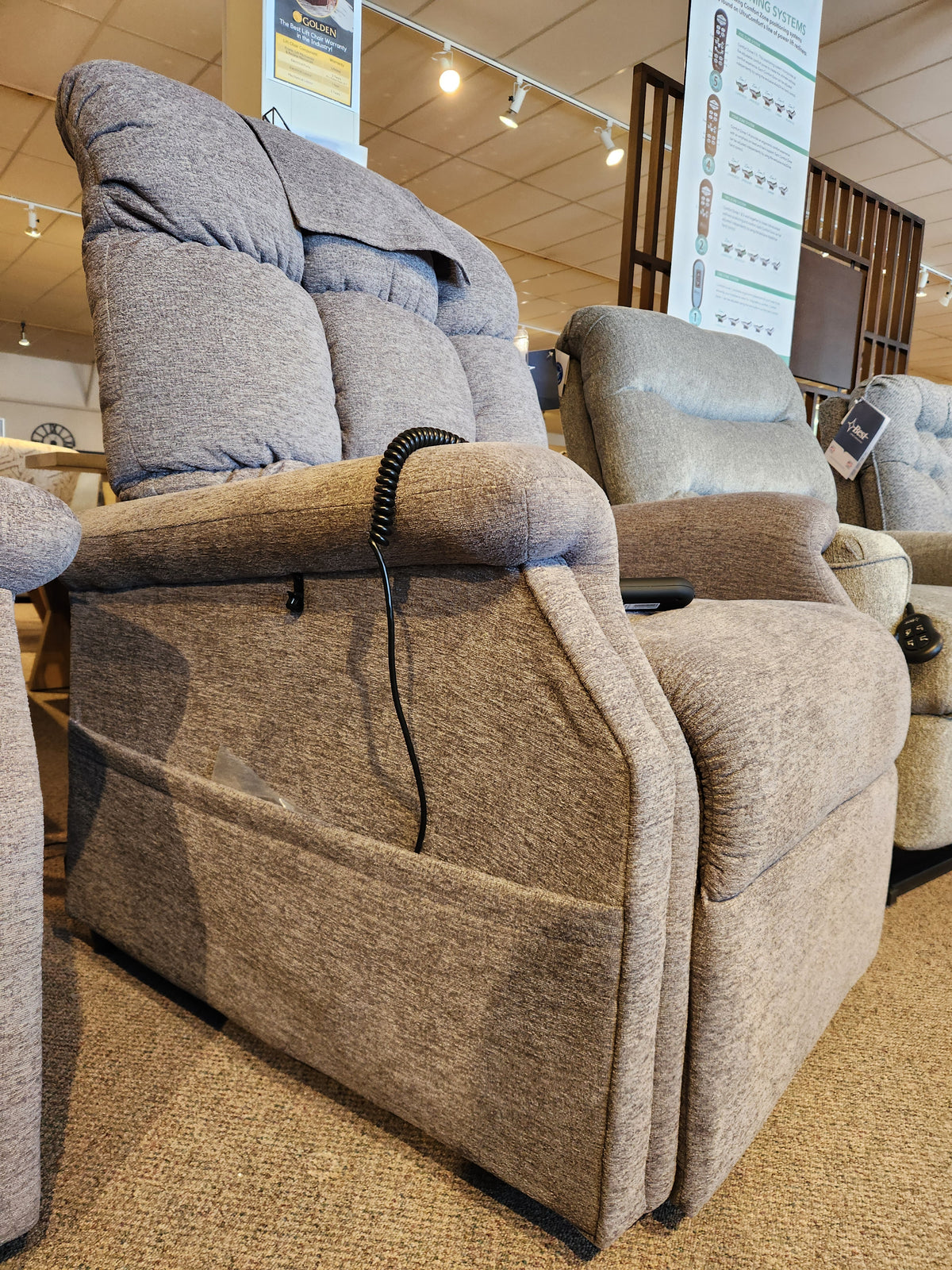 A Golden Aurora PL480 beige upholstered power lift chair with remote is on display in a furniture showroom.