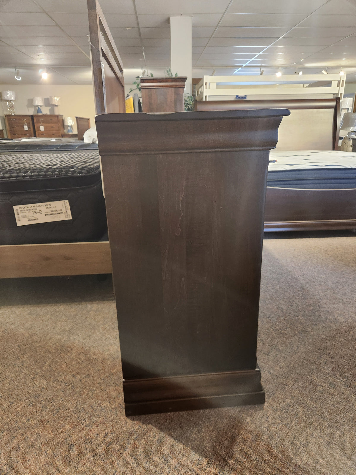 The Vokes 260 Louis Rustique 6 Drawer Dresser, a dark wood piece, is on display with beds and mattresses in-store.