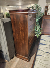 The Vokes 260 Louis Rustique 5 Drawer Chest in dark wood stands tall in a showroom beside a bed and plant.