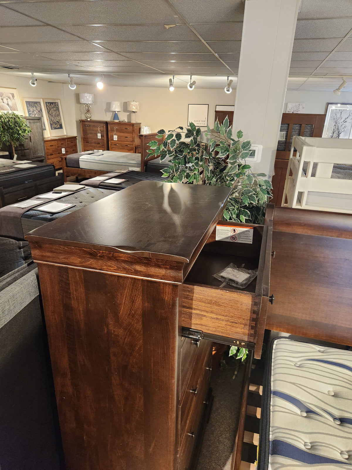 A Vokes 260 Louis Rustique 5 Drawer Chest is displayed with an open drawer among beds and decor in a showroom.