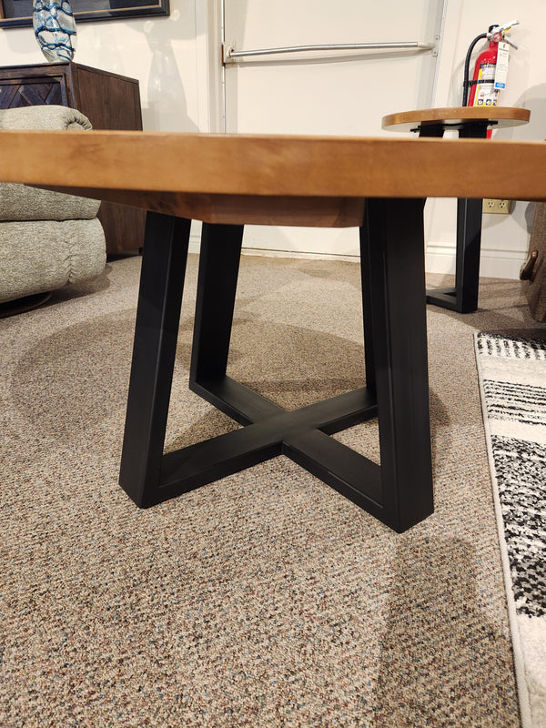 The Handstone Belmont Round Coffee Table with a black metal X-base stands on a carpeted floor among modern furniture.