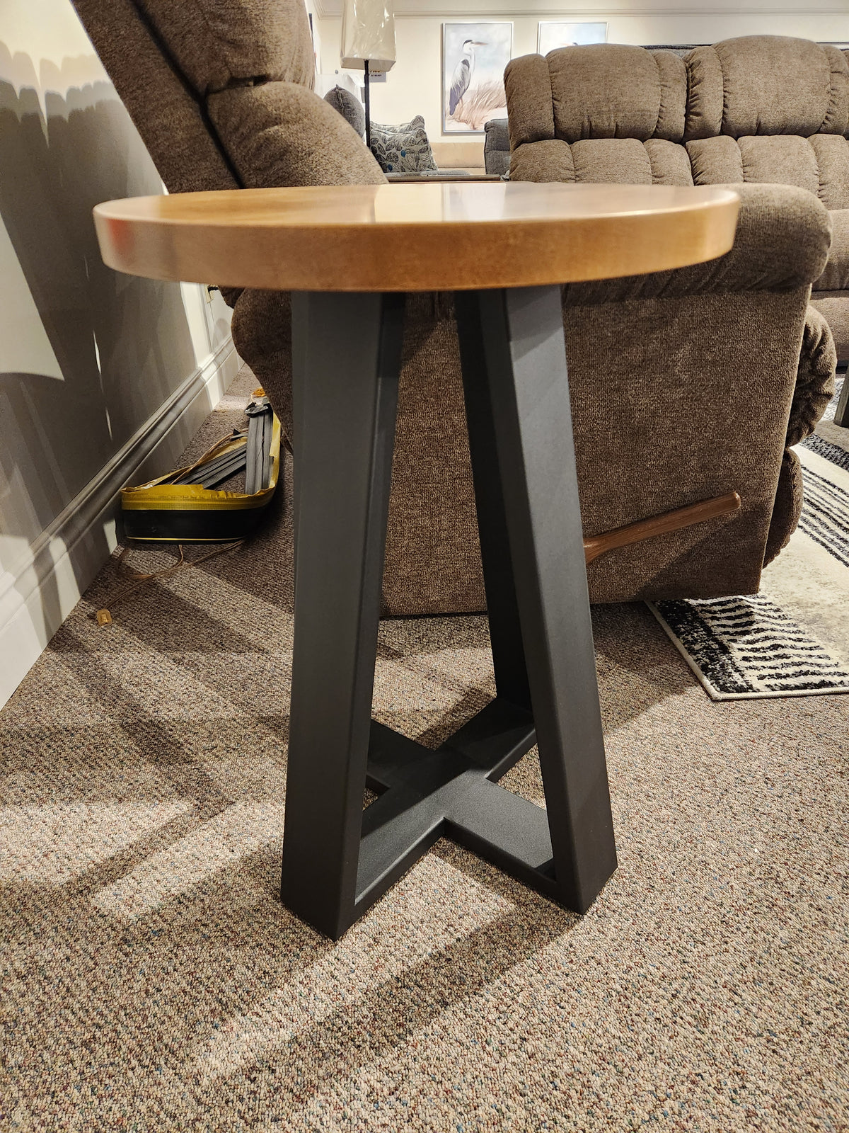 The Handstone Belmont Round Chairside Table with black metal legs stands on a carpeted floor next to a recliner.