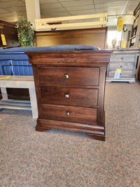 The Vokes 260 Louis Rustique 3 Drawer Nightstand is displayed amid rustic bedroom furniture in the store.
