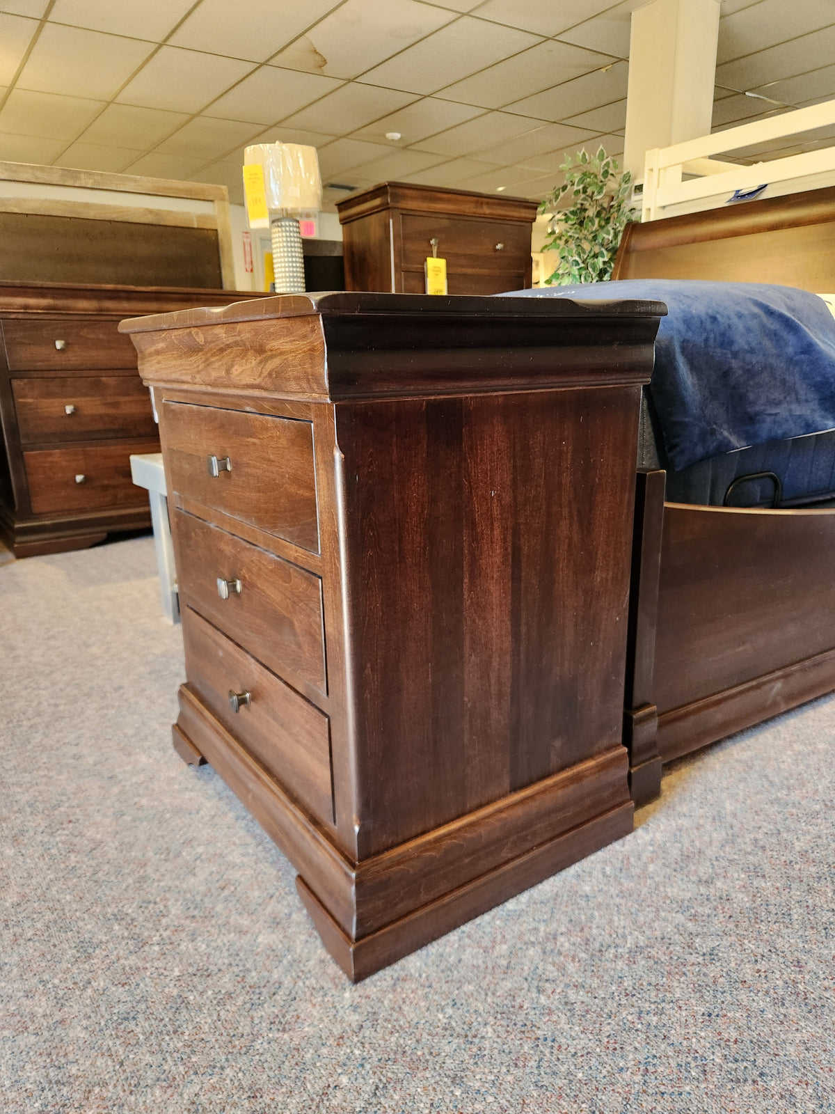 The Vokes 260 Louis Rustique 3 Drawer Nightstand sits on carpet in a showroom with other rustic bedroom pieces.