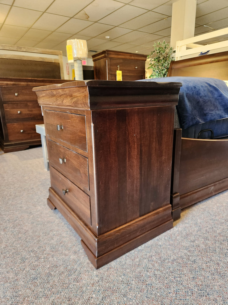 The Vokes 260 Louis Rustique 3 Drawer Nightstand sits on carpet in a showroom with other rustic bedroom pieces.