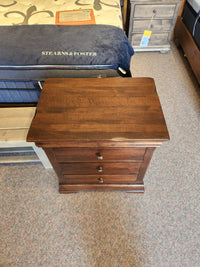 A Vokes 260 Louis Rustique 3 Drawer Nightstand in wormy maple stands beside a mattress and another nightstand.