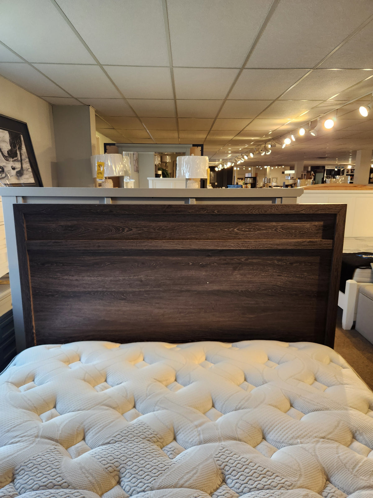 The Magnussen B6308 Merrifield Queen Storage Bed is displayed with a beige mattress under bright showroom lighting.