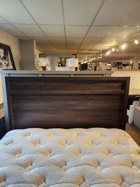 The Magnussen B6308 Merrifield Queen Storage Bed is displayed with a beige mattress under bright showroom lighting.