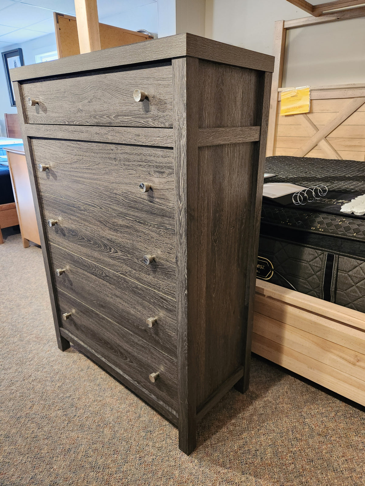 A Magnussen B6308 Merrifield Chest in dark oak is displayed next to a mattress and bed frame in the showroom.