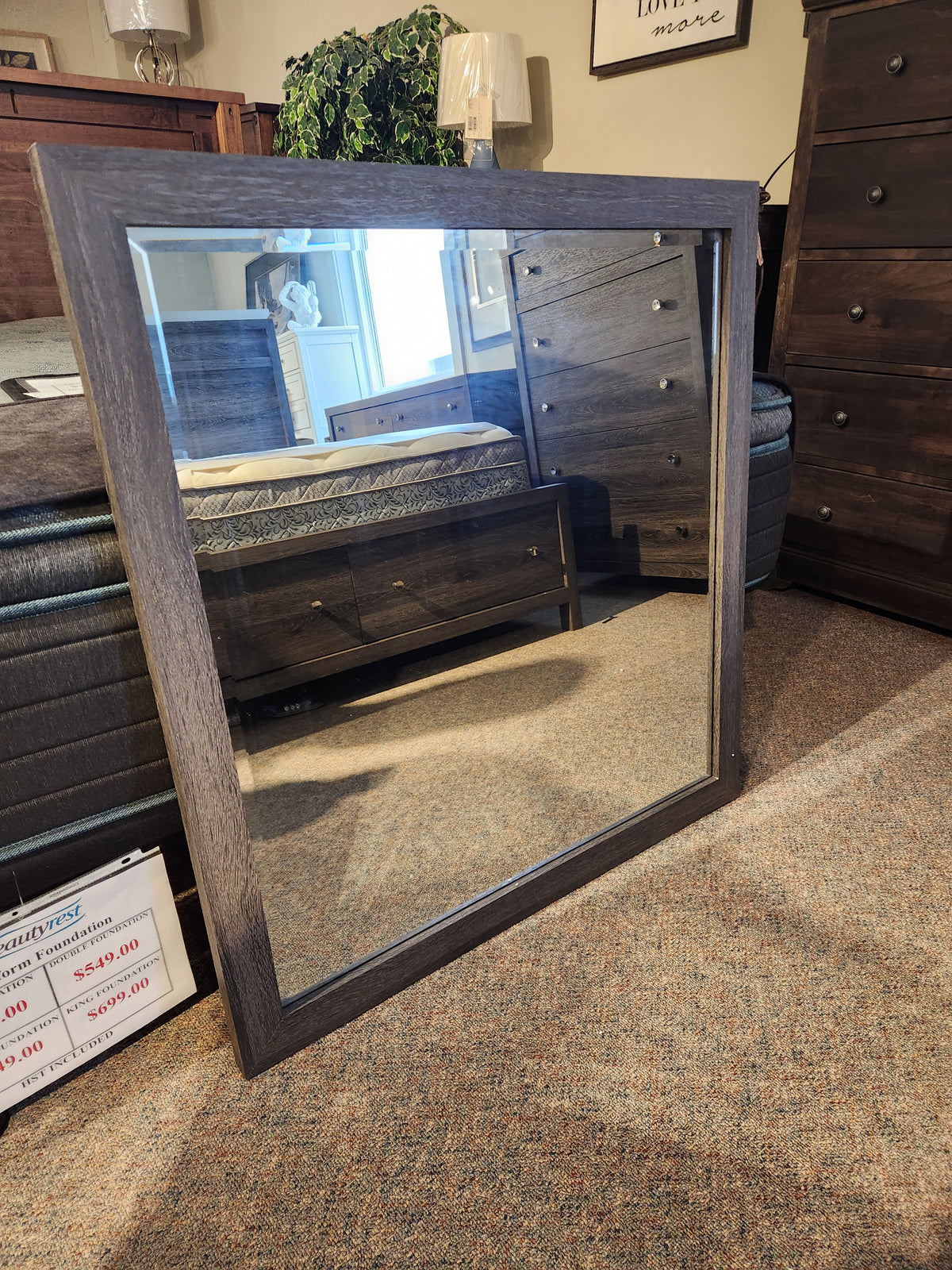 The Magnussen B6308 Merrifield Mirror with a wooden frame rests on the floor, reflecting modern bedroom furniture.
