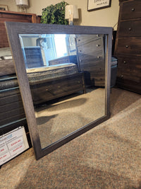 The Magnussen B6308 Merrifield Mirror with a wooden frame rests on the floor, reflecting modern bedroom furniture.