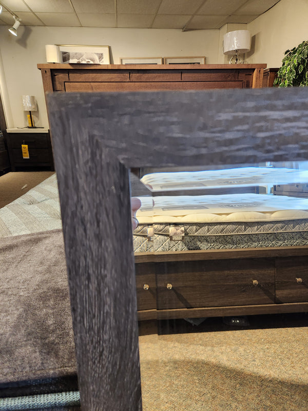 The B6308 - Merrifield Mirror by Magnussen reflects a wooden bed frame and mattress in a modern bedroom setting.