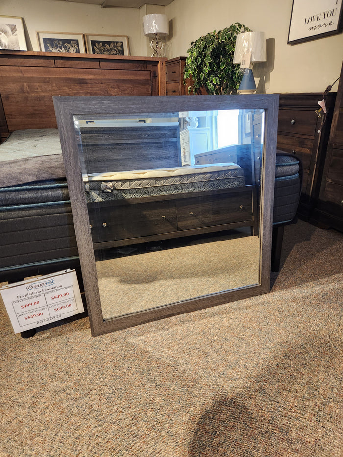 The Magnussen B6308 - Merrifield Mirror with oak veneer frame rests on the bedroom showroom’s carpeted floor.