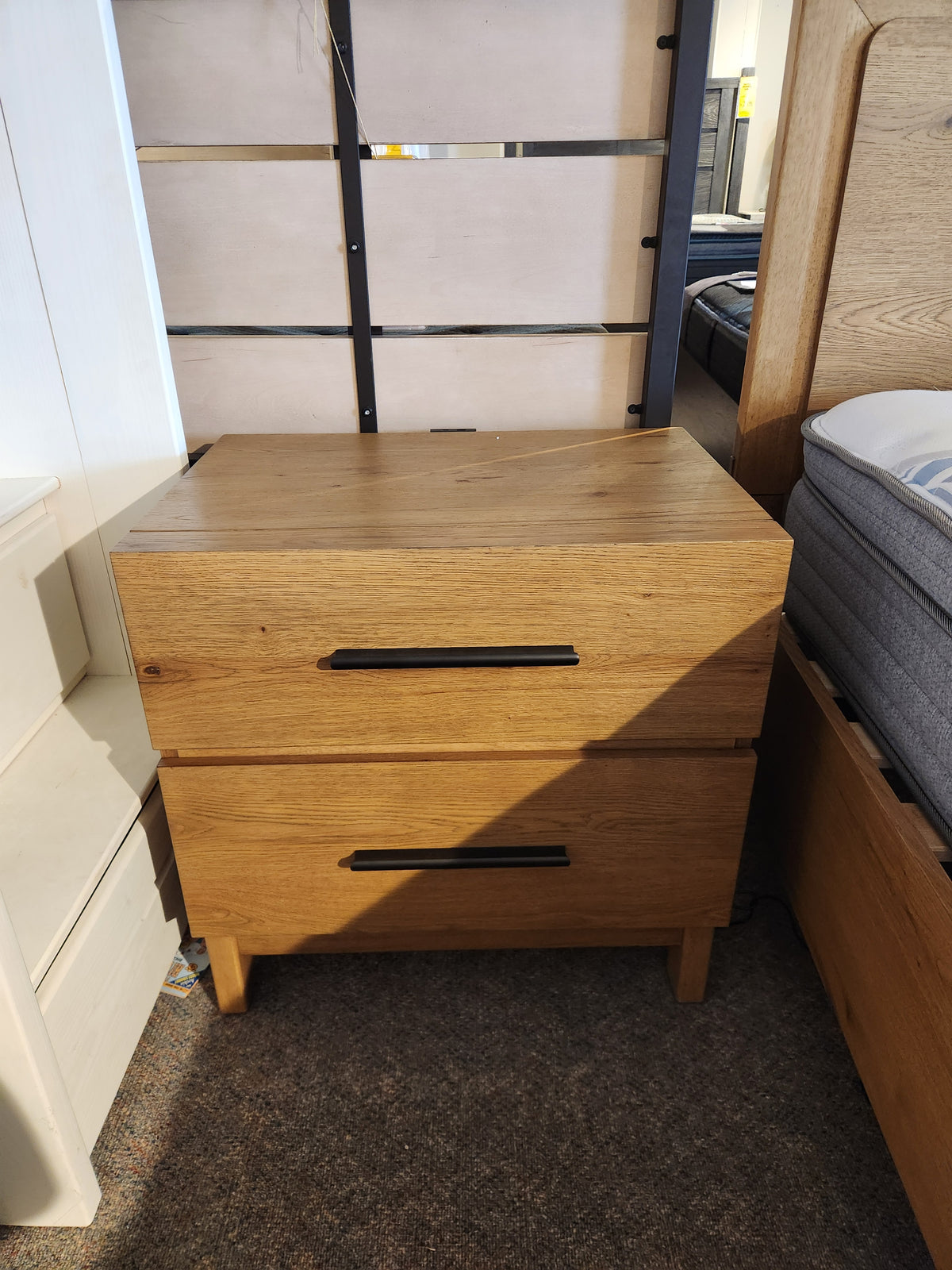The Magnussen B6309 Davenport Nightstand, rustic caramel, 2 drawers, sits on carpet between store beds.