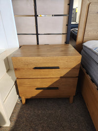 The Magnussen B6309 Davenport Nightstand, rustic caramel, 2 drawers, sits on carpet between store beds.