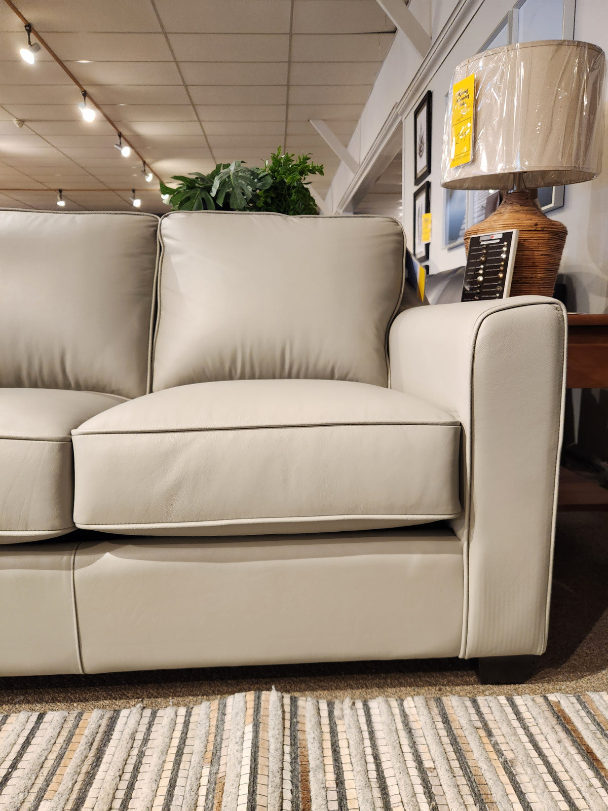 A Leather Craft Sophia Sofa in light gray is beside a side table with a lamp wrapped in plastic on carpeted flooring.