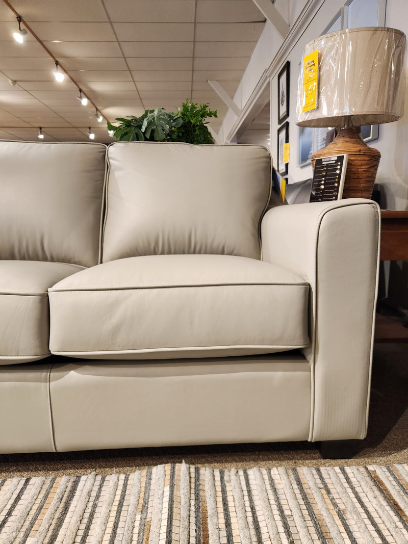 A Leather Craft Sophia Sofa in light gray is beside a side table with a lamp wrapped in plastic on carpeted flooring.