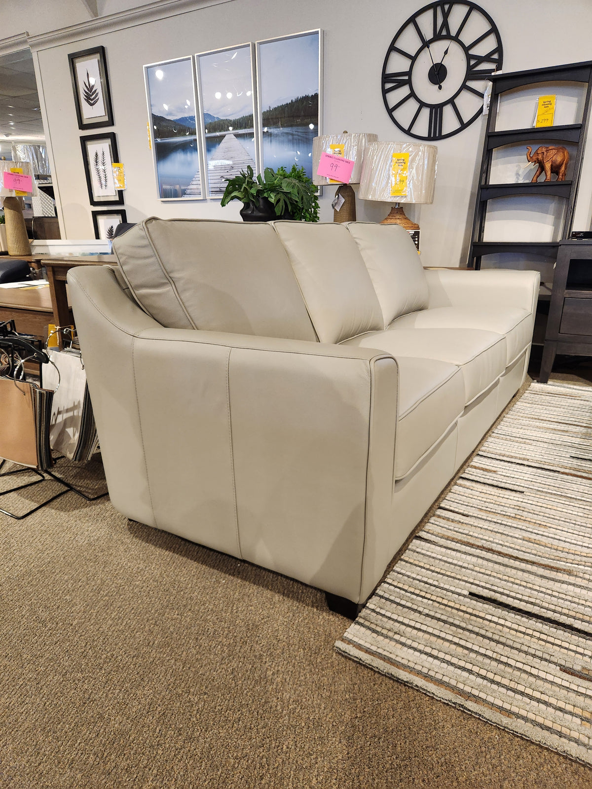 The Leather Craft Sophia Sofa is shown in a showroom with wall art, a clock, and shelves in the background.