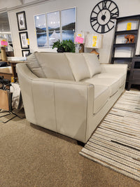 The Leather Craft Sophia Sofa is shown in a showroom with wall art, a clock, and shelves in the background.