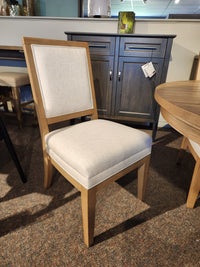 A Magnussen D6480-Coventry wooden chair in Honey Maple with beige upholstery is displayed on the showroom floor.