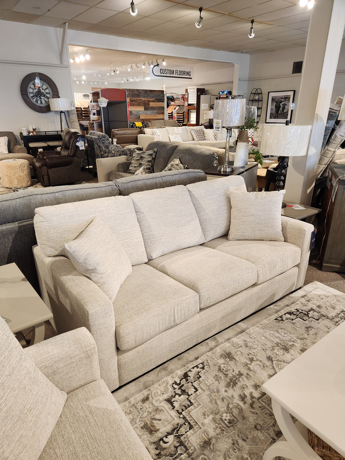 The Superstyle 9540 Stationary Sofa, beige with cushions, is shown in a store among other sofas and decor.