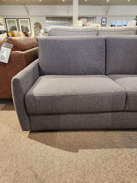 The Primo Vitale Sofa Bed in gray upholstery with memory foam is showcased on a beige carpet in the showroom.