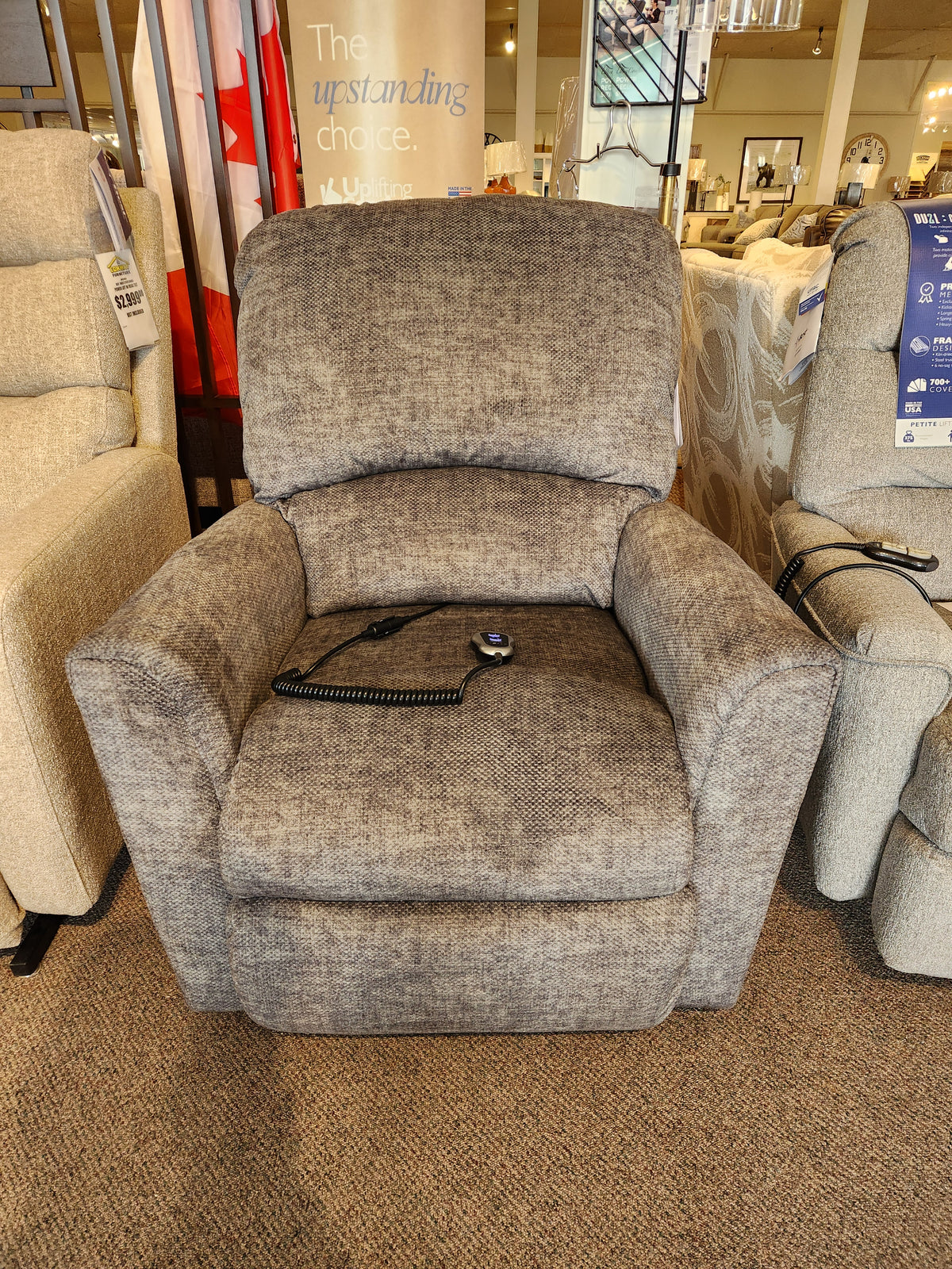 The La-z-boy 793 Jean Bronze Lift Chair with wired remote sits on a carpeted showroom floor between other chairs.
