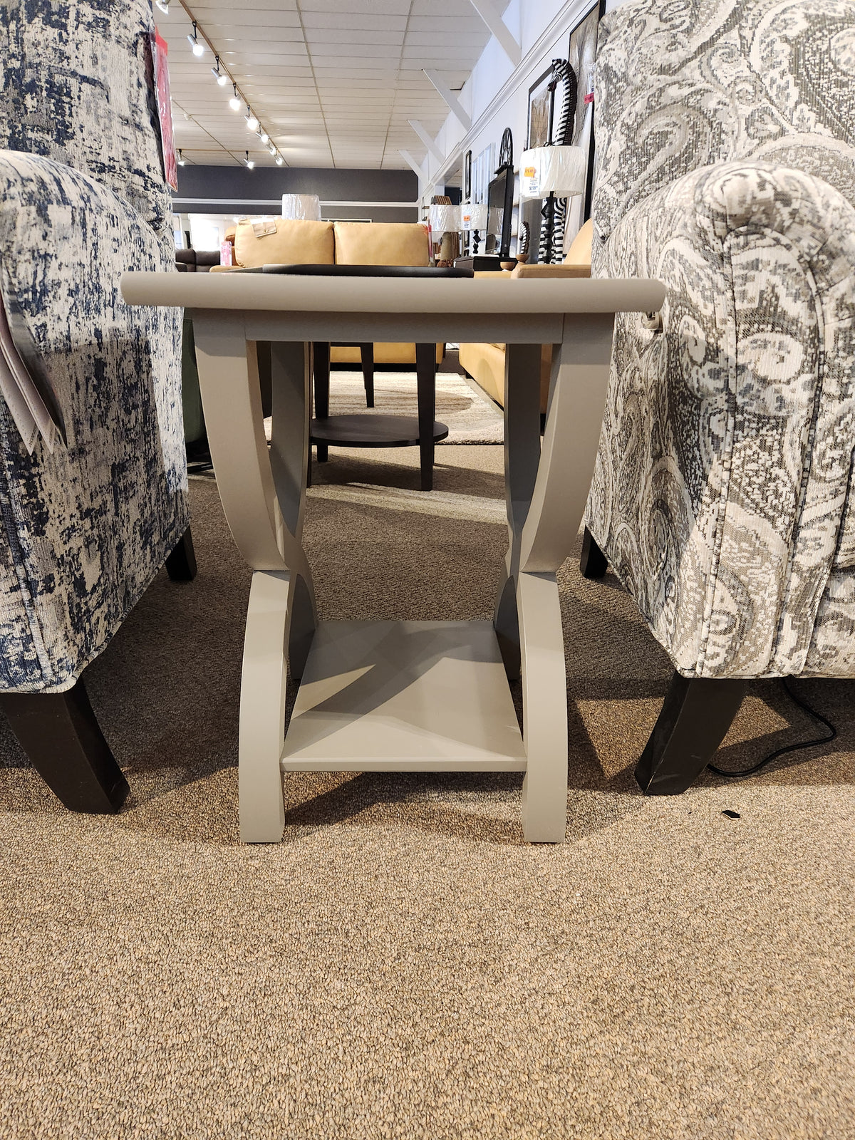 The Durham 900-532 Transitional Chairside End Table with curved legs sits between two armchairs on a carpeted floor.