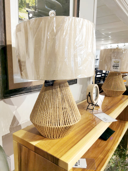 Ashley Clayman Table Lamp with a woven base and beige lampshade on display in a store.