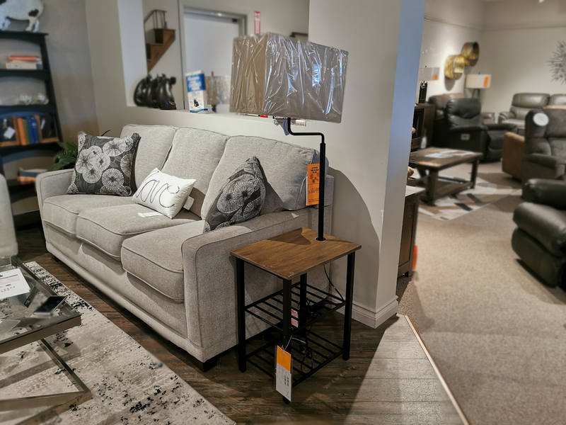 A light gray sofa with patterned pillows sits by a wooden side table and Ashley’s Shianne Floor Lamp in a showroom.