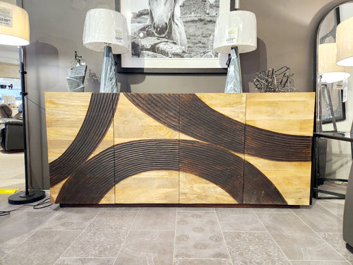 The Crestview Collection 4593 Lennox 4 Door Sideboard with curved patterns stands between two lamps in a modern showroom.