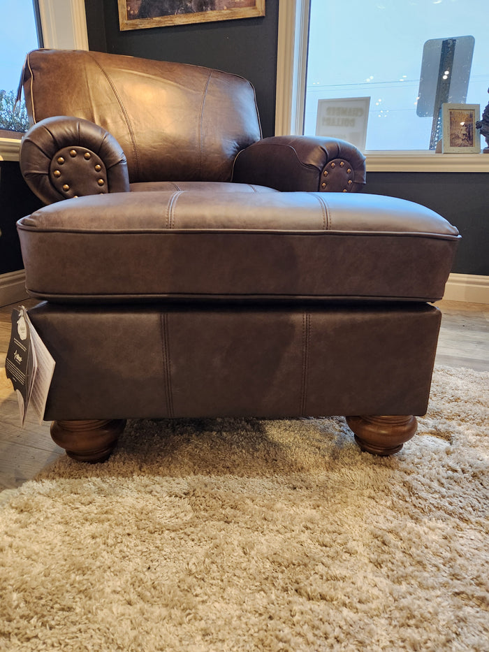 The Best F24ELU Noble Leather Ottoman in brown rests on a beige shag rug in a room with large windows.