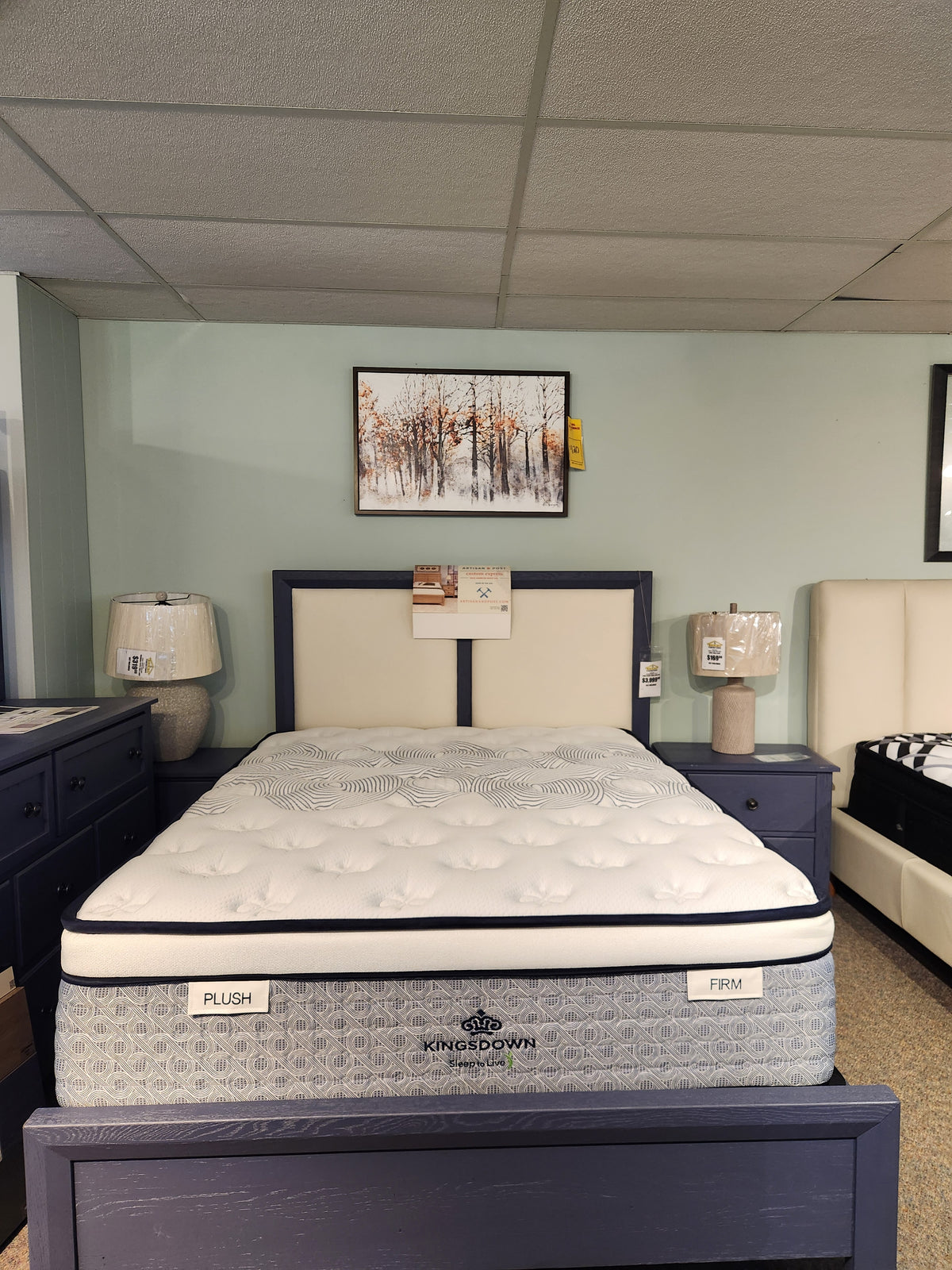 A showroom display featuring the KingsDown Radiance Elite 2 ET Firm Mattress, flanked by nightstands and art above.