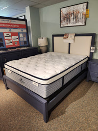 A KingsDown Radiance Elite 2 ET Firm queen mattress on a blue frame, with nightstands, lamp, and wall art in showroom.