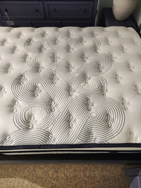 A close-up of the KingsDown Radiance Elite 2 ET Firm Mattress with geometric stitching; navy blue dresser in background.