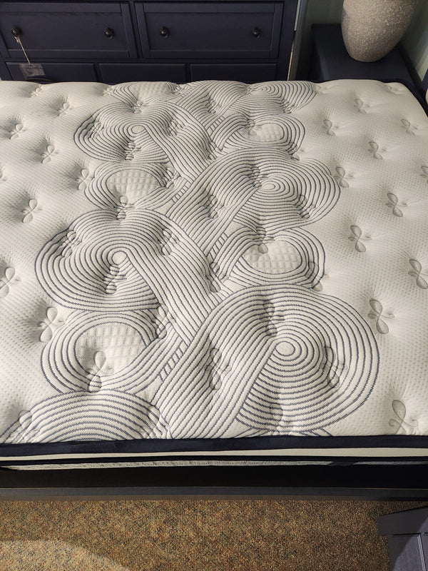A close-up of the KingsDown Radiance Elite 2 ET Firm Mattress with geometric stitching; navy blue dresser in background.