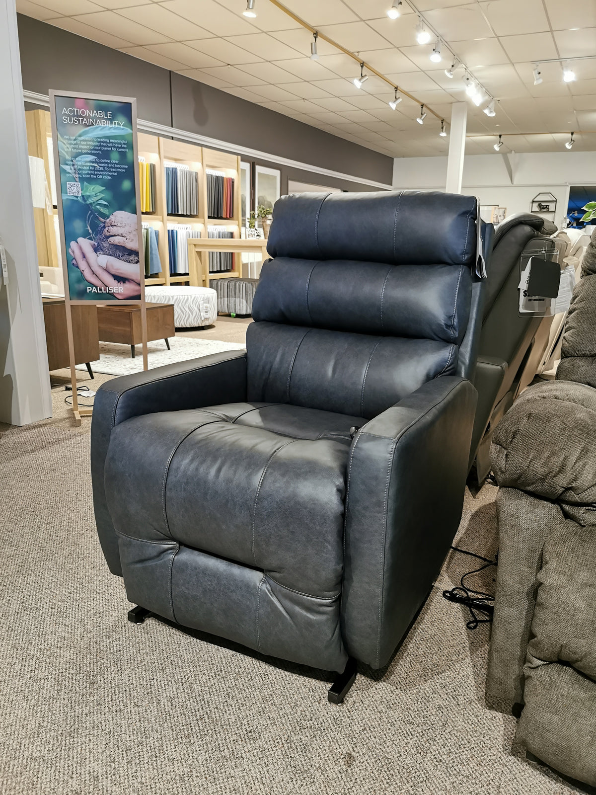 The Kristisha 9NZ31LV Power Lift Chair by Best, in dark gray leather, is showcased on a carpeted showroom floor.