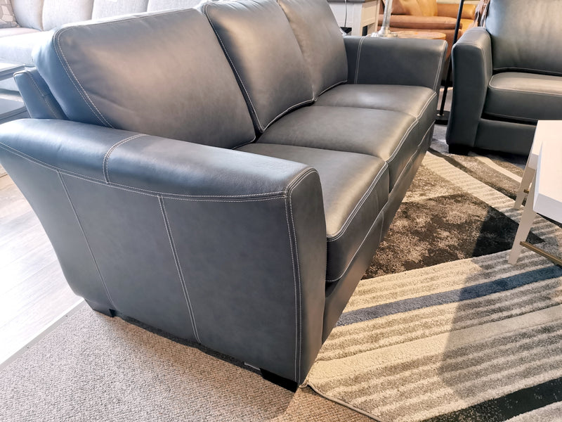 The Bayview Leather Sofa by Leather Craft, upholstered in gray leather, sits elegantly on a textured rug in the showroom, blending traditional charm with contemporary style amid an array of other furniture pieces.
