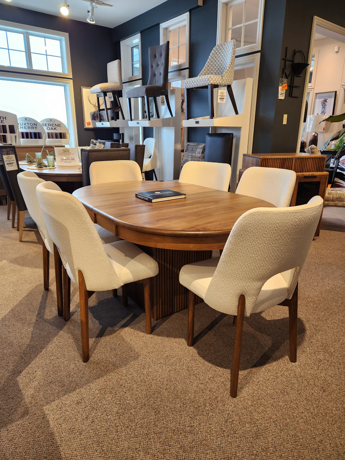 Handstone Lotus Dining Package: Wooden pedestal table with 6 cushioned wood chairs in a modern showroom.