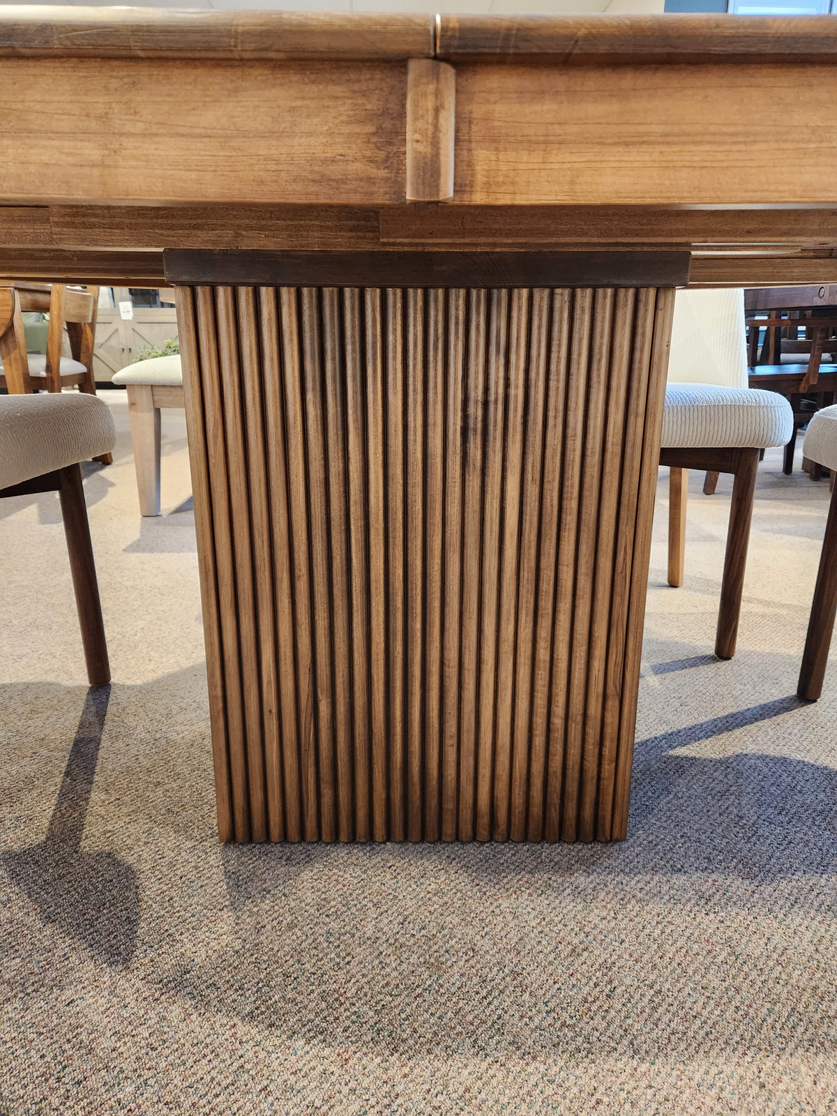 Close-up of a Lotus Dining Package table leg by Handstone, paired with solid wood chairs on a carpeted floor.