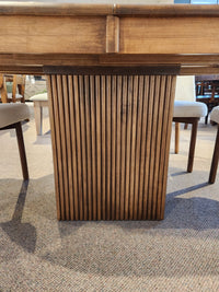 Close-up of a Lotus Dining Package table leg by Handstone, paired with solid wood chairs on a carpeted floor.