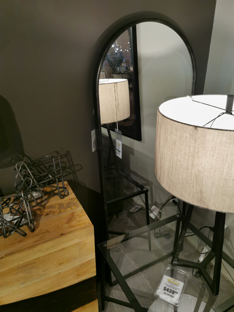 The Mercana 68739 Agatha Full Length Arc Mirror stands behind a lamp on a glass table by a wooden dresser in-store.