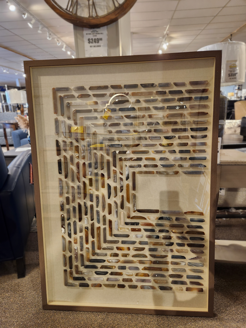 A framed art piece titled "70298 Stone Trails Agate II" by Mercana, featuring a geometric brick pattern and dimensional texture, displayed on the floor in a store setting with the price tag visible above.