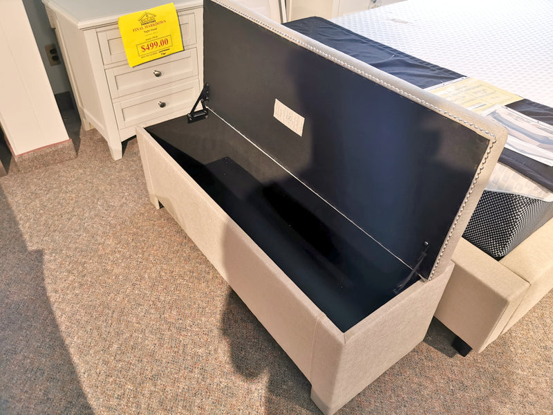 A beige Titus Upholstered Storage Bench with its lid open sits by a white nightstand in a furniture store.