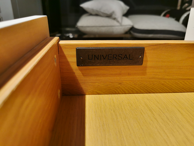 A wooden drawer from the Universal 987350 Summerhill 3 Drawer Night Table, "UNIVERSAL" label inside, cushions behind.